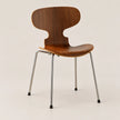 A mid-century modern wooden chair with a curved backrest and seat made of warm brown wood, supported by slim chrome metal legs with black feet, set against a plain beige background.