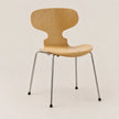 A modern wooden dining chair with a light natural oak seat and backrest, supported by slim chrome metal legs with black rubber feet, set against a plain beige background.