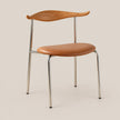 A mid-century modern dining chair with a curved light brown wood backrest, a tan leather seat cushion, and a chrome metal frame, shown against a solid beige background.