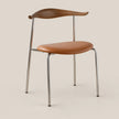 A mid-century modern dining chair featuring a curved wooden backrest, a smooth tan leather seat, and slim chrome legs, set against a solid beige background.