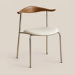A modern dining chair with a curved wooden backrest, a cream upholstered seat, and slim metallic legs, shown against a solid beige background.