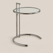 A modern round side table with a clear glass top and a polished chrome metal base, shown against a solid beige background.