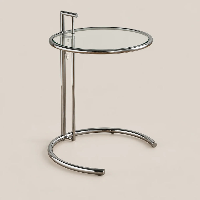 A modern round side table with a clear glass top and a polished chrome metal base, shown against a solid beige background.