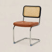 A modern side chair with a tufted brown leather seat, a black-framed cane backrest, and a chrome cantilever steel base, shown against a solid light beige background.