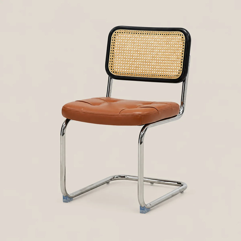 A modern side chair with a tufted brown leather seat, a black-framed cane backrest, and a chrome cantilever steel base, shown against a solid light beige background.
