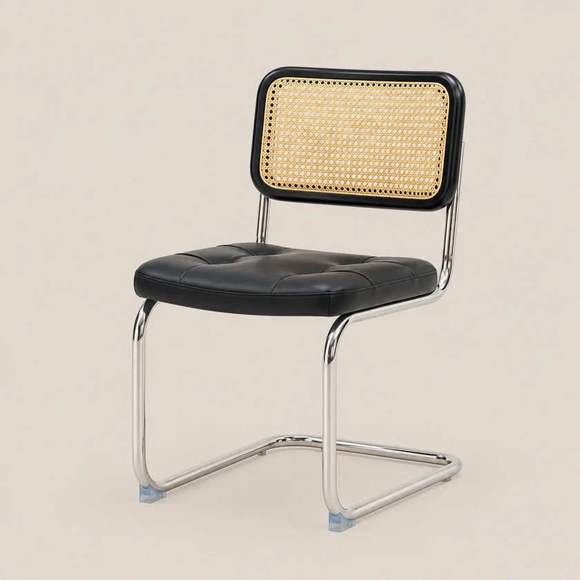 A modern cantilever dining chair with a black faux leather seat and a woven cane backrest, supported by a polished chrome metal frame, set against a solid light beige background.