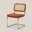 A modern side chair with a tufted cognac brown leather seat and a woven cane backrest, supported by a chrome cantilever frame on a light beige background.