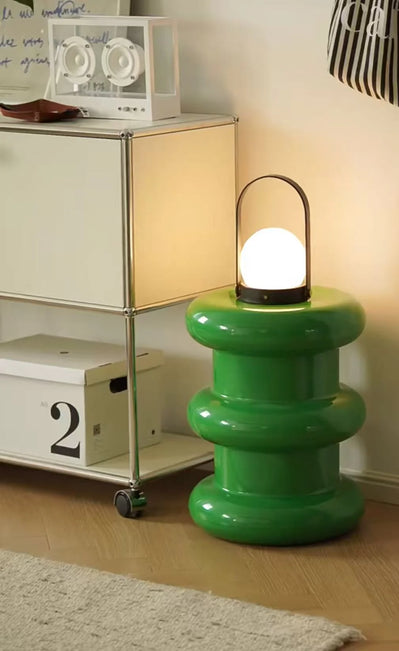 Green lamp with a white bulb on a wooden floor, next to a metal shelf.