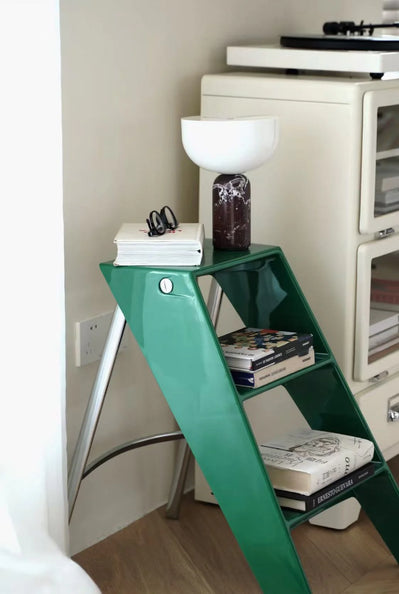 Green step ladder used as a shelf with books and a lamp in a room.
