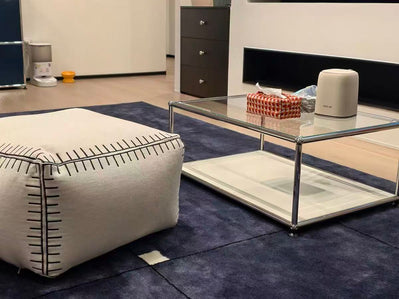 Modern living room with a glass coffee table, ottoman, and decorative items.