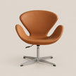 A modern swivel lounge chair with a warm tan leather shell and a silver metal four-star base, photographed against a solid light beige background.