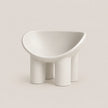A minimalist white molded chair with a wide, rounded seat and four cylindrical legs, photographed on a clean, solid warm beige background.