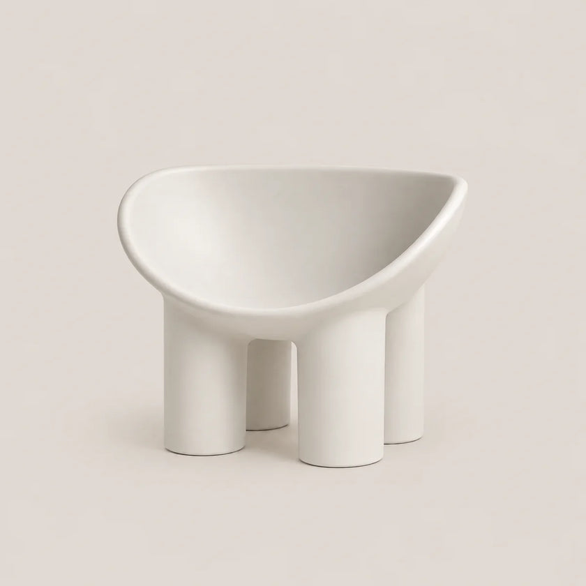 A minimalist white molded chair with a wide, rounded seat and four cylindrical legs, photographed on a clean, solid warm beige background.