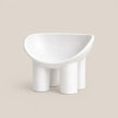 A minimalist white molded chair with a wide, rounded seat and four cylindrical legs, shown against a solid beige background.