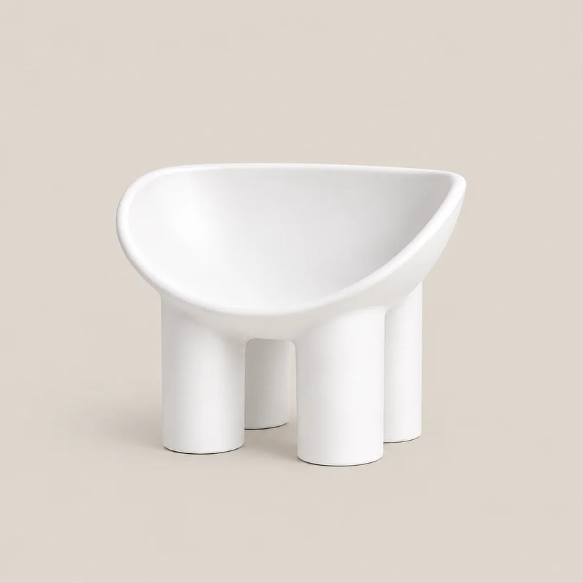 A minimalist white molded chair with a wide, rounded seat and four cylindrical legs, shown against a solid beige background.