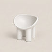 A minimalist white molded chair with a wide, rounded seat and four cylindrical legs, photographed on a clean, solid warm beige background.