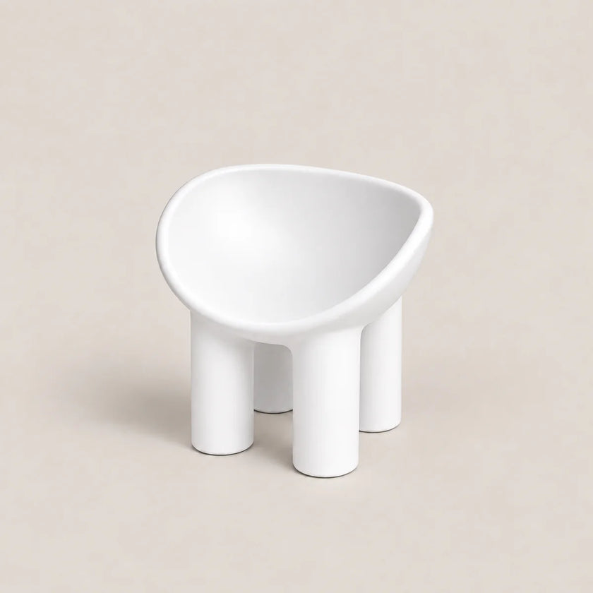 A minimalist white molded chair with a rounded, bowl-like seat and four cylindrical legs, set against a solid beige background.