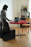 Person using a laptop on a red table in a room with a shelf and curtains.