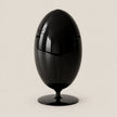 A glossy black egg-shaped decorative object with a pedestal base, photographed against a plain warm light beige background.
