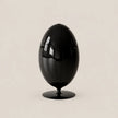 A glossy black egg-shaped sculpture or decorative object on a matching round base, photographed against a plain warm light beige studio background.
