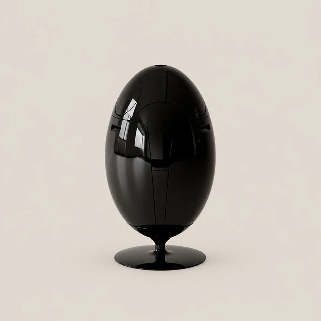 A glossy black egg-shaped sculpture or decorative object on a matching round base, photographed against a plain warm light beige studio background.