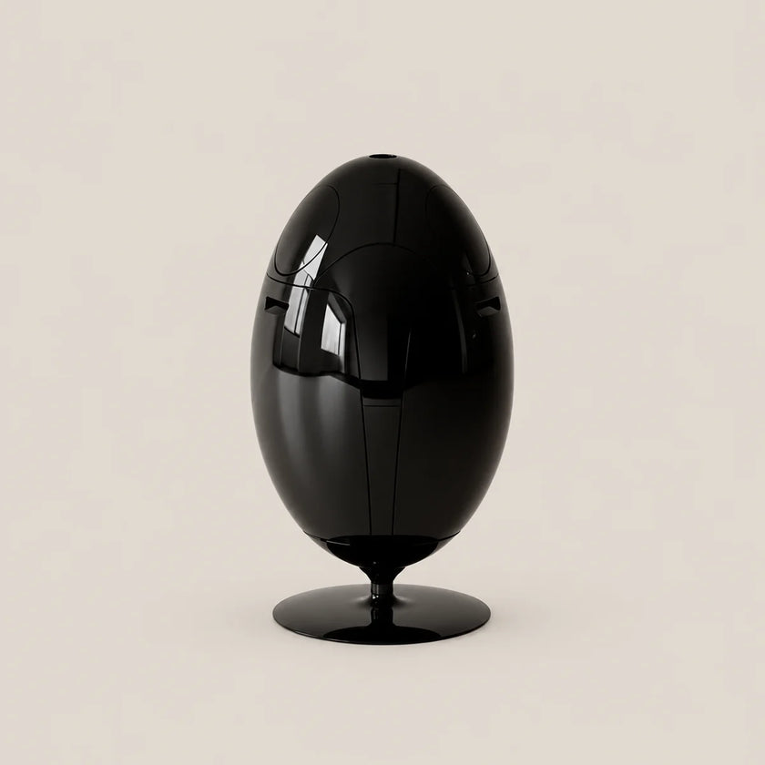 A glossy black egg-shaped sculpture or decorative object on a matching round base, photographed against a plain warm light beige studio background.