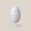 A white egg-shaped countertop appliance with a circular top panel and a small pedestal base, shown centered on a plain warm beige background.