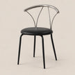 A modern dining chair with a black upholstered seat and black legs, featuring a curved polished silver metal backrest, set against a solid light beige background.