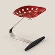 A modern red perforated seat with a curved chrome support base and a black footrest, photographed on a plain warm beige studio background.