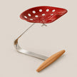 A modern industrial-style stool with a glossy red seat featuring white polka dots, supported by a curved polished steel base and a light natural wood footrest, set against a solid beige background.