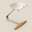 A modern industrial-style stool with a white perforated plastic seat, a curved polished metal base, and a light brown wooden footrest, shown on a solid light beige background.