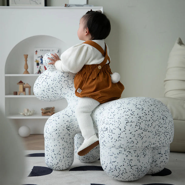 Child sitting on a speckled ottoman in a room with shelves and decor.