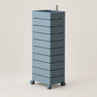 A tall, modern rolling storage unit with nine stacked rectangular drawers in a muted blue-gray color, featuring a square top with a sink-like recess and a small metal handle, set against a solid light beige background.