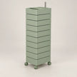 A tall, narrow rolling storage cart with multiple stacked drawers in a muted sage green color, featuring a metal handle on top and small caster wheels at the base, shown against a plain light gray studio background.