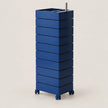 A tall, blue rolling organizer with multiple stacked drawers and a top tray holding a metal utensil, photographed on a clean light beige studio background.
