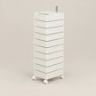 A tall, narrow white rolling storage cabinet with multiple drawers and a small metal handle on top, shown on a plain warm beige studio background.