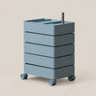 A modern four-drawer rolling storage cart in a muted dusty blue-gray color, featuring a silver metal handle on top and four caster wheels, set against a solid light beige background.