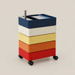 A stackable rolling storage cart with five stacked rectangular drawers or bins in navy, cream, yellow, orange, and red, on black wheels with a metal handle.