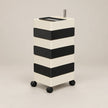 A modern rolling storage cart with four black-and-cream horizontal drawers and black caster wheels, shown on a plain warm light-gray studio background.