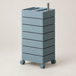 A tall, modular rolling storage cart with six stacked drawer-like compartments, finished in a muted dusty blue-gray color, featuring a silver metal handle on top and caster wheels at the base, set against a plain beige studio background.