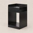 A minimalist black metal side table with a rectangular top tray, two open shelves, and a lower base, shown on a solid light beige background.