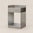 A minimalist, open-frame side table or shelf made of brushed stainless steel, featuring a top tray, a central open cubby, and a bottom shelf, set against a solid light beige background.