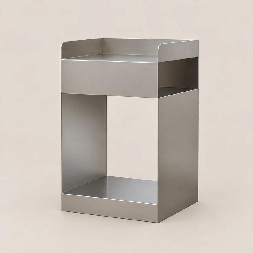 A minimalist, open-frame side table or shelf made of brushed stainless steel, featuring a top tray, a central open cubby, and a bottom shelf, set against a solid light beige background.