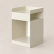 A minimalist, modern side table or organizer with a cream-colored finish, featuring a top tray, a middle open shelf, and a lower base shelf, set against a solid light beige background.