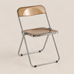 A modern folding chair with a translucent smoky brown seat and backrest, supported by a slim chrome metal frame, shown on a solid light beige background.
