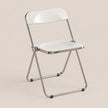 A modern folding chair with a glossy white seat and backrest, supported by a slim metallic silver frame, set against a solid light beige background.
