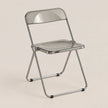A modern folding chair with a transparent smoky-gray seat and backrest, supported by a polished silver metal frame, set against a solid light beige background.