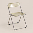 A modern folding chair with a transparent, frosted pale yellow seat and backrest, supported by a polished chrome metal frame, set against a solid beige background.