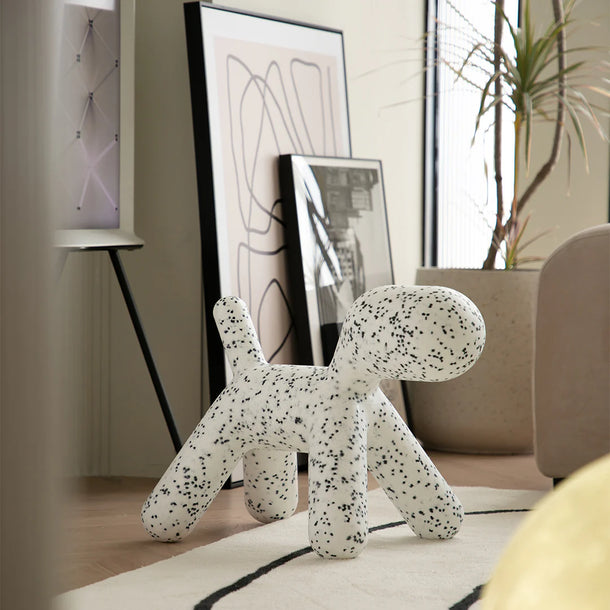 Speckled dog-shaped object on a surface with abstract art and a plant in the background