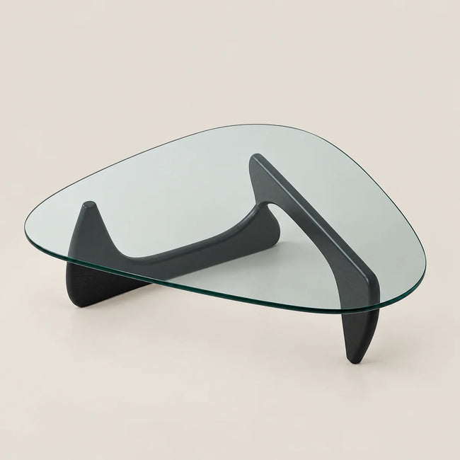 A modern glass-top coffee table with a dark charcoal curved sculptural base, photographed on a clean, solid, warm off-white background.